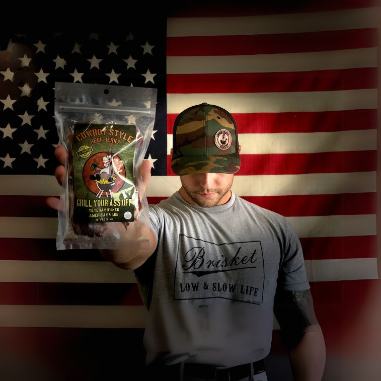 A man holds a clear package of Grill Your Ass Off Cowboy Style Beef Jerky in front of an American flag backdrop.