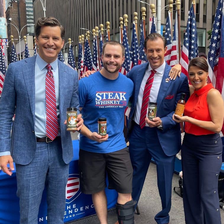 A photo featuring Grill Your Ass Off's Steak and Whiskey spices being showcased on a television segment, with the American flag visible in the background.