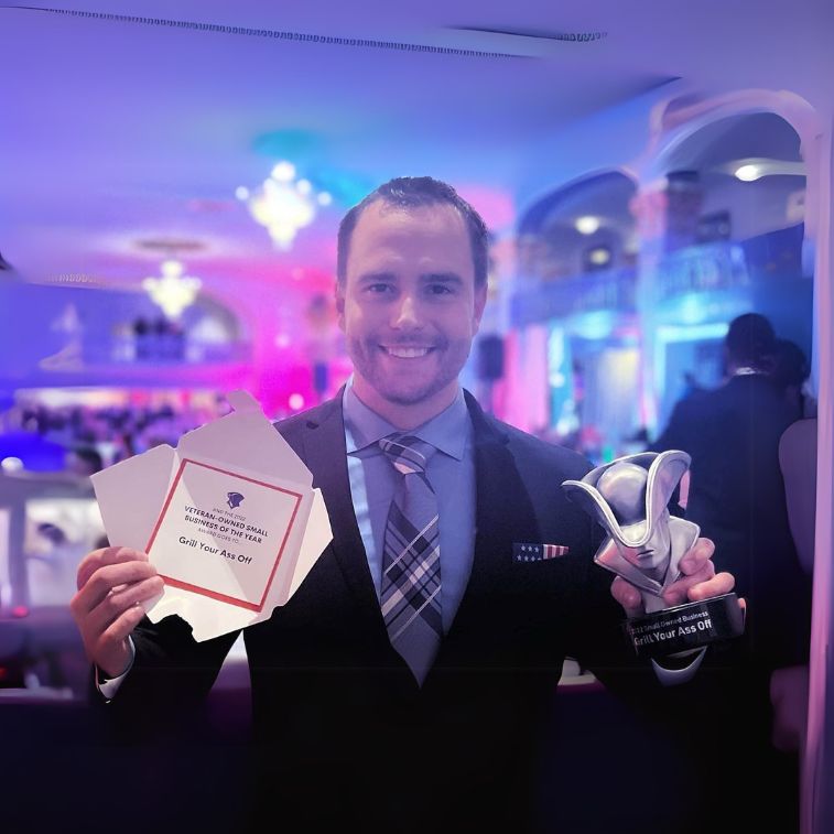 Jason, the owner of Grill Your Ass Off, smiles while holding the "Veteran-Owned Small Business of the Year" award and the accompanying certificate.